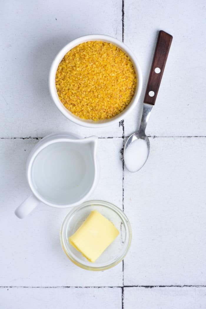 A bowl of bulgar, a pitcher of water, and a glass dish with butter sit ready on the white tiled background, along with a spoon of salt—essential elements mirroring the simplicity found in learning how to cook bulgur wheat.
