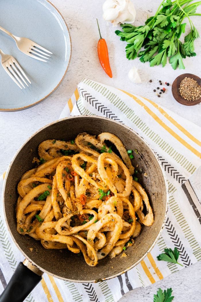 A pan of fried calamari with herbs and spices rests on a striped cloth beside forks, garlic, pepper, and parsley, much like an inviting setting for learning how to cook calamari.