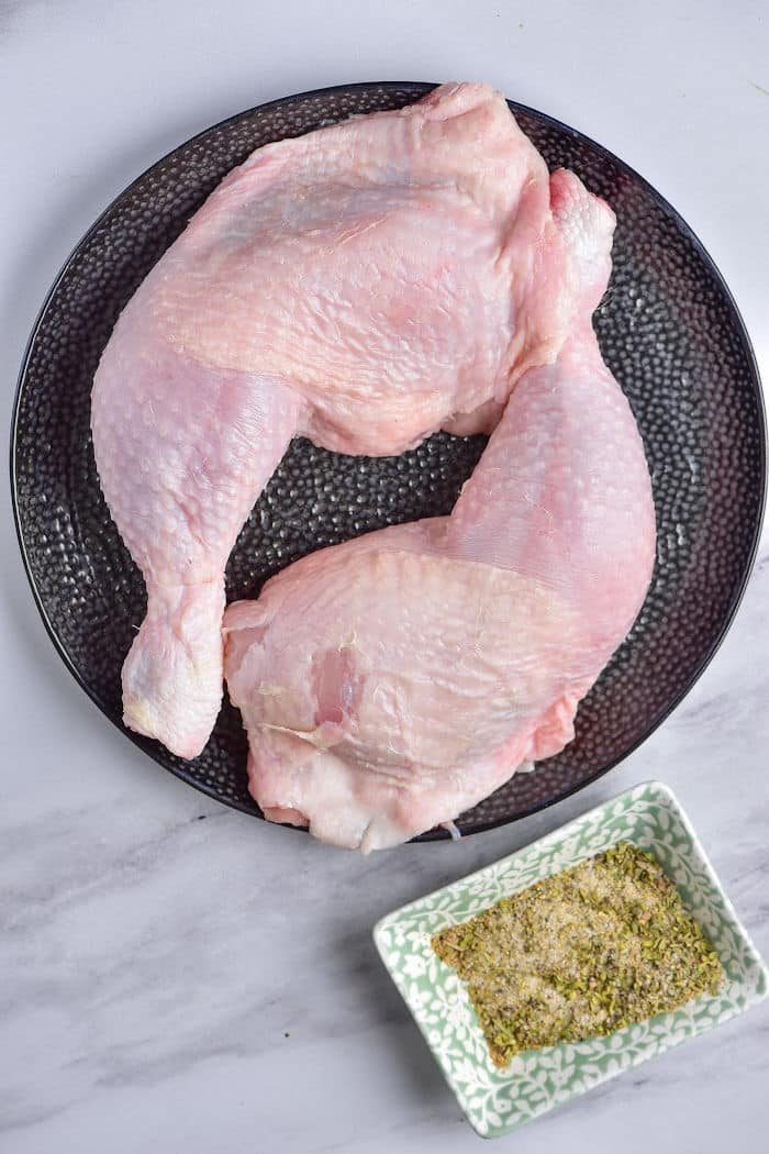 Raw chicken quarters rest invitingly on a black plate, accompanied by a small dish of spices on the marble surface—your first step in mastering how to cook chicken leg quarters to perfection.
