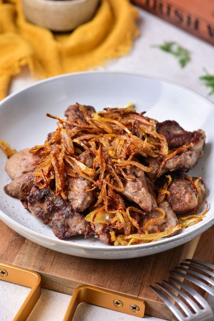 A plate of grilled chicken liver topped with crispy fried onions sits on the table, tempting you to pick up the nearby fork. This dish perfectly complements a guide on how to cook chicken liver to perfection.