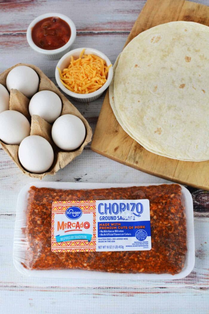 A wooden surface displays tortillas on a cutting board, eggs in a carton, grated cheddar cheese in a bowl, salsa in a small bowl, and a package of Kroger Mercado ground chorizo sausage—solid ingredients for learning how to cook chorizo and eggs.