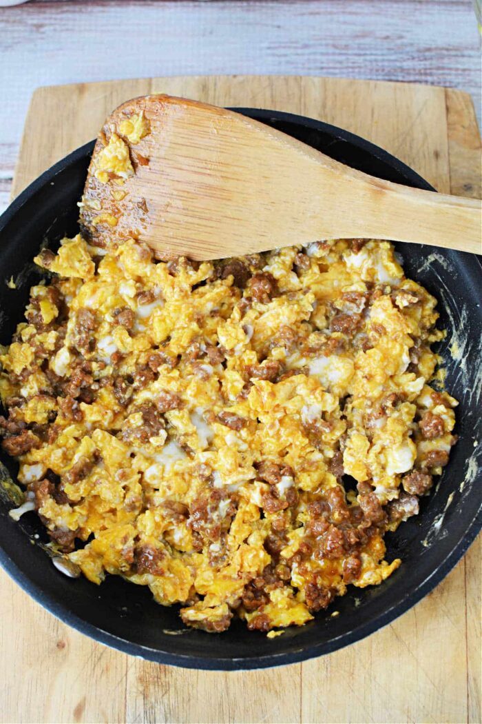 A frying pan on a wooden cutting board contains scrambled eggs mixed with ground sausage. A wooden spatula rests on the edge of the pan, partially covered in the egg and sausage mixture—a solid demonstration of how to cook chorizo and eggs.