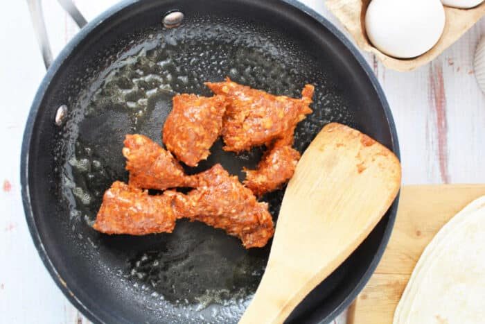 A black skillet sizzles with pieces of chorizo being cooked, a wooden spatula resting in the pan. Nearby on the counter, a carton of eggs awaits its turn, next to a tortilla on a wooden cutting board—solid for learning how to cook chorizo and eggs.