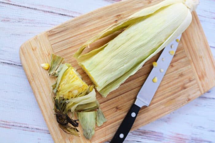 A partially peeled corn cob with husk rests alongside a knife on a wooden cutting board, ready for an adventure in oven cooking.