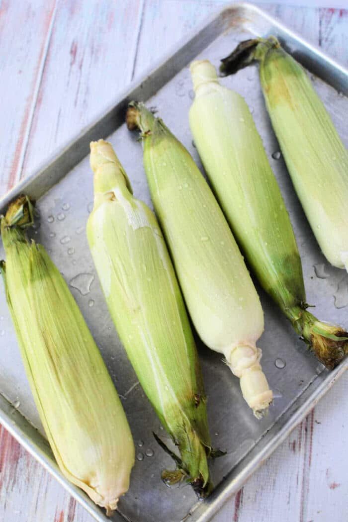 Five ears of corn in husks rest on a metal baking sheet, some glistening with water droplets—solid for learning how to cook corn on the cob in the oven on a white wooden surface.