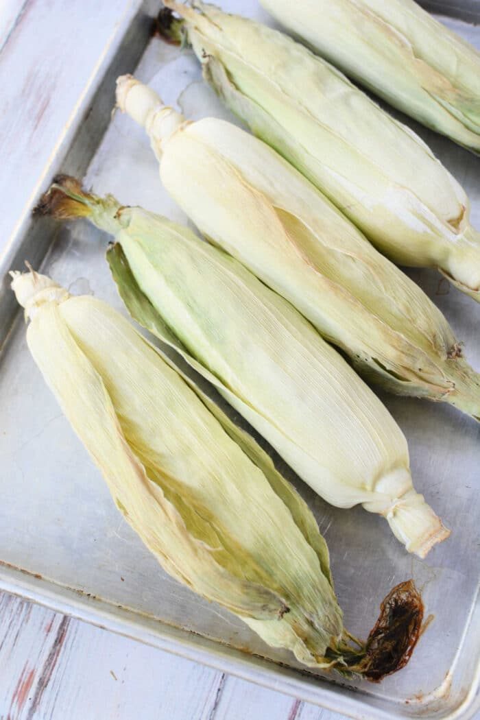 Five unpeeled ears of corn on the cob rest side by side on a metal baking tray, ready for oven cooking, arranged neatly on a wooden surface.