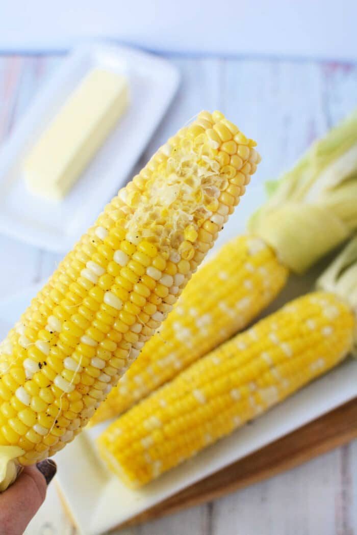 A close-up shows a buttered ear of corn on the cob, with two unbuttered ones and a stick of butter in the background, capturing the essence of oven-cooked perfection.