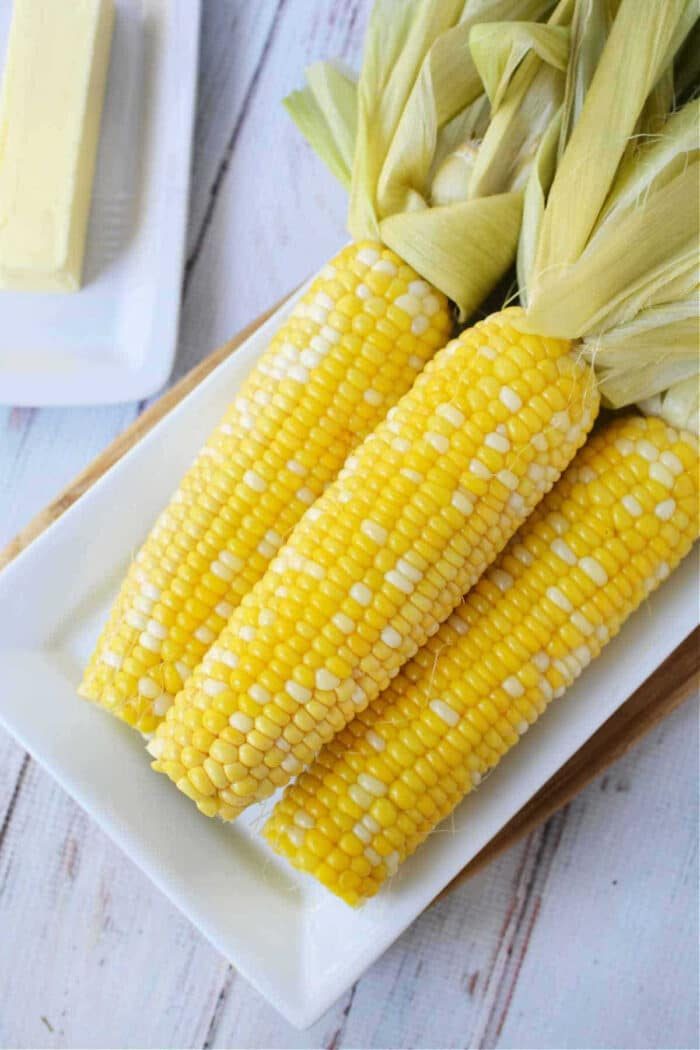 Four ears of fresh corn on the cob with husks, placed on a white rectangular plate—solid for learning how to cook corn on the cob in the oven for a simple and good side dish.
