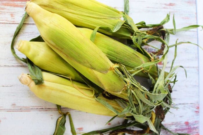 Fresh ears of corn with husks and leaves rest on a wooden surface, ready for oven cooking.