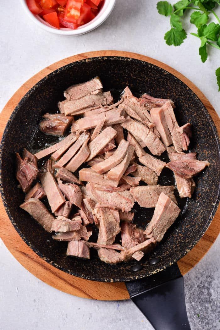 Sliced cooked tongue in a frying pan on a wooden board, next to cut tomatoes and cilantro. For those interested in expanding their culinary skills, learning how to cook cow tongue can offer a rich and delectable addition to your kitchen repertoire.