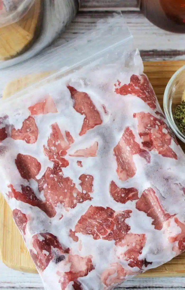 A clear plastic bag holds frozen pieces of red meat, solid for those wondering how to cook frozen beef. On a wooden cutting board beside the bag is a small bowl with green herbs, resting on what seems to be a kitchen countertop.