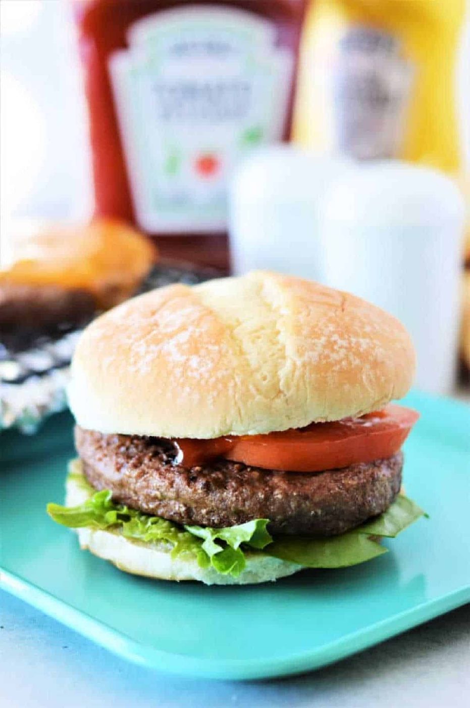 A hamburger with lettuce and tomato on a blue plate, with ketchup and mustard bottles in the background—solid inspiration for learning how to cook frozen burgers in the oven for a quick, good meal.