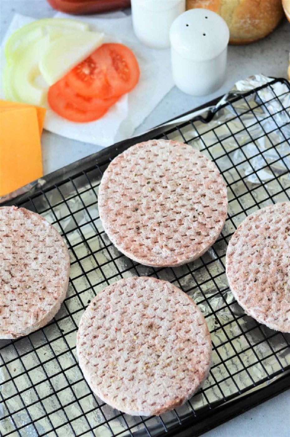 Four frozen hamburger patties on a wire rack, with sliced tomato, onion, cheese, and seasonings nearby—solid for learning how to cook frozen burgers in the oven for a quick and tasty meal.