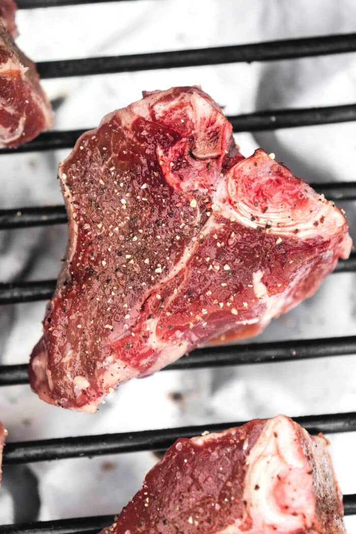 A raw piece of seasoned lamb loin chop sits on a grill, showcasing visible fat marbling and a sprinkle of black pepper and other spices. For those learning how to cook lamb loin chops, the background teases another piece of meat partially visible on the metal grill.
