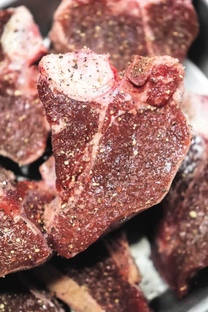 Close-up of raw, seasoned lamb loin chops piled together. The meat is seasoned with visible black pepper and salt, similar to the methods used in How to Cook Lamb Loin Chops. The texture of the steaks is marbled with some fat and dark red lean meat.