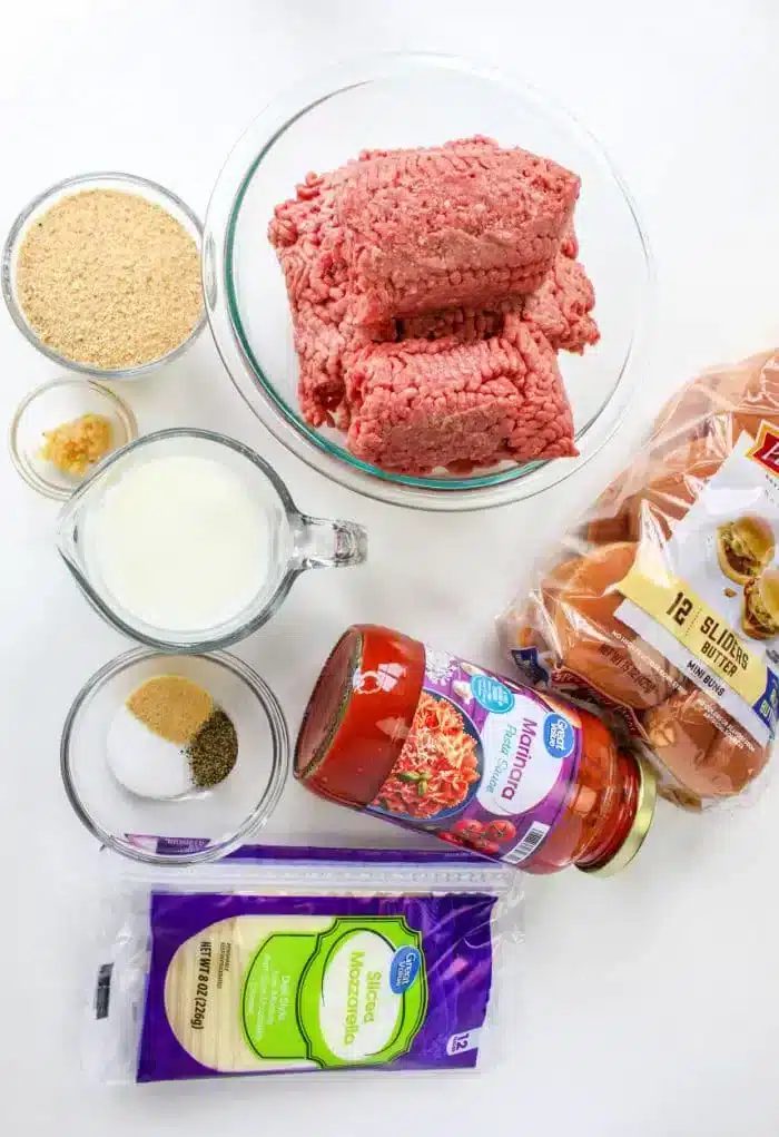 On a white surface, ground beef rests beside slider buns, marinara sauce, and sliced mozzarella cheese. Accompanied by breadcrumbs and small bowls of minced garlic and seasoning, a cup of milk completes the setup for learning how to cook meatballs on the stovetop.