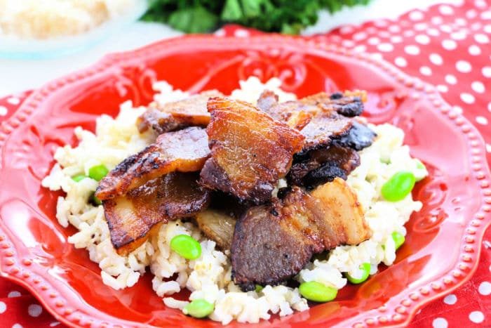 Crispy bacon pieces—solid for those learning how to cook pork belly in the oven—are served on a bed of rice with green edamame on a red plate.