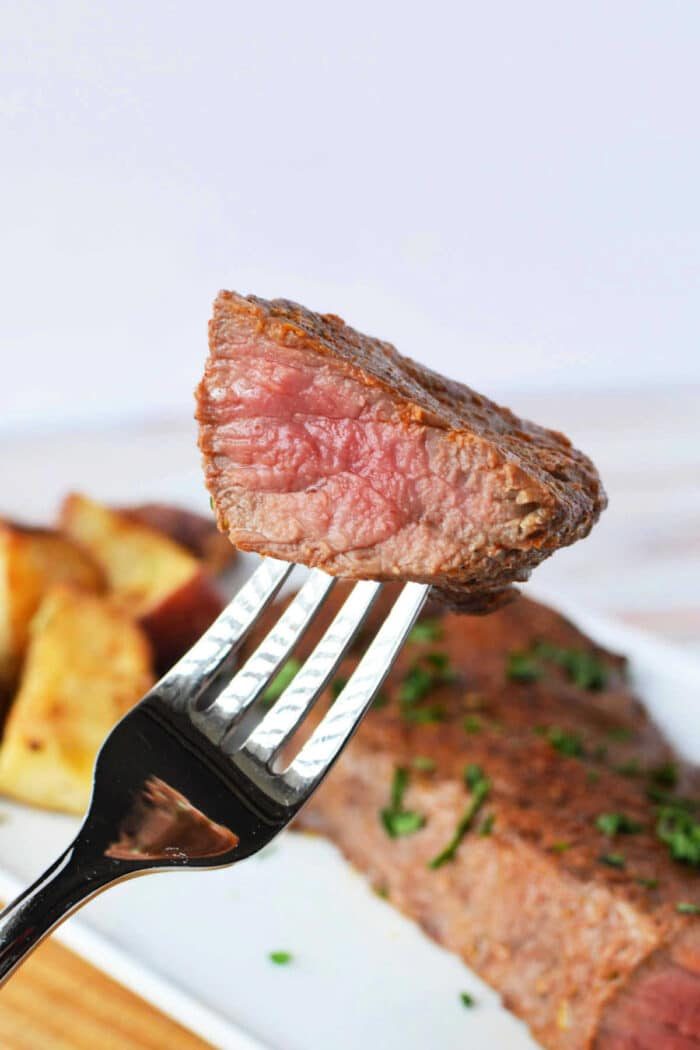 A fork holds a piece of juicy, medium-rare tri tip steak above a plate featuring steak and roasted potatoes. For those wondering how to cook tri-tip in the oven, this succulent meal showcases the solid harmony of tender meat and crispy sides.