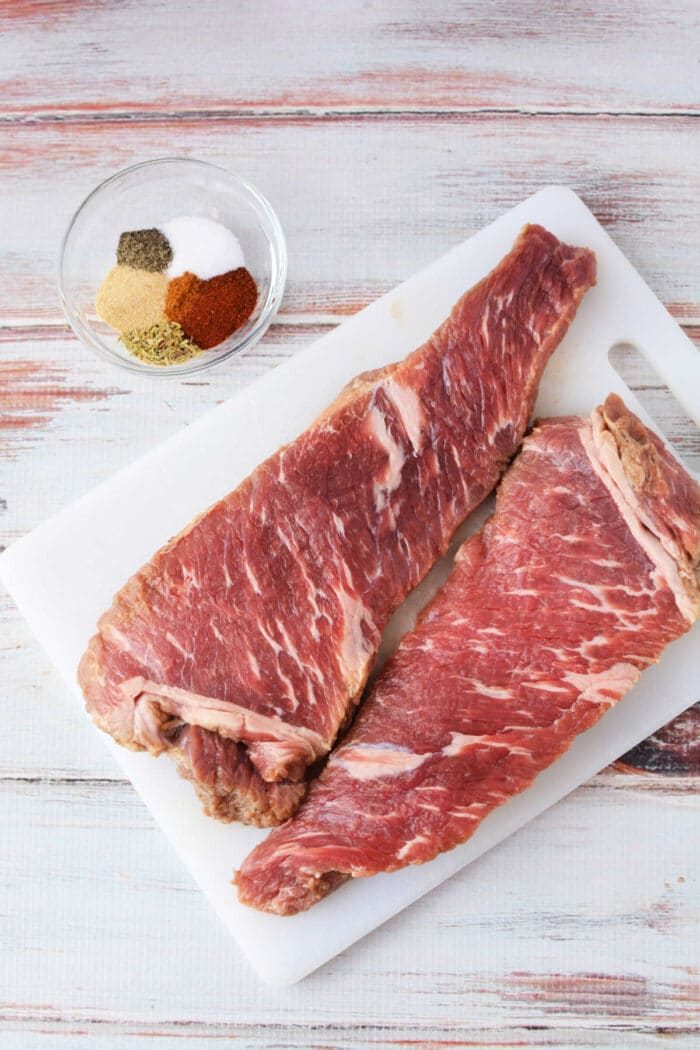 Two tri tip raw steaks sit on a cutting board alongside a bowl of spices, ready to transform into a mouthwatering dish. solid for enthusiasts figuring out how to cook tri tip in the oven, this setup promises savory success on any wooden surface.