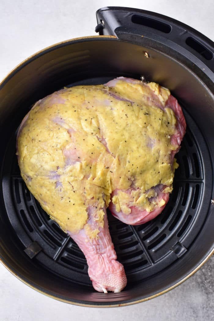 A raw turkey leg, perfectly coated with a yellow marinade, sits in an air fryer basket, ready to show you how to cook turkey legs and thighs to perfection.