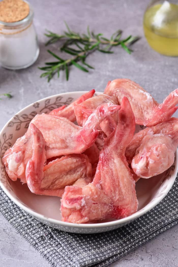 A bowl of raw chicken wings sits on a gray kitchen cloth adorned with fresh rosemary, oil, and a jar of salt in the background. Discover how to defrost chicken in the microwave to prepare your feast quickly and efficiently.
