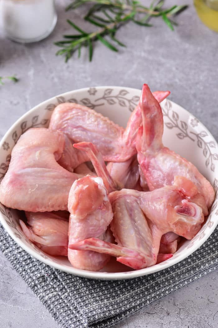 A bowl of raw chicken wings on a gray textured surface, garnished with fresh herbs in the background, prepped and ready for you to learn how to defrost chicken in the microwave efficiently.