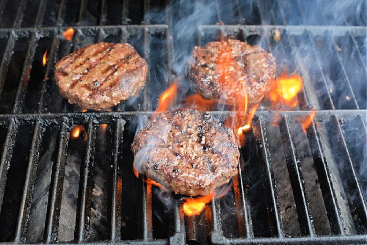 Three beef patties grilling over open flames and smoke on a barbecue grill—discover how to grill frozen burgers to perfection with this mouthwatering scene.