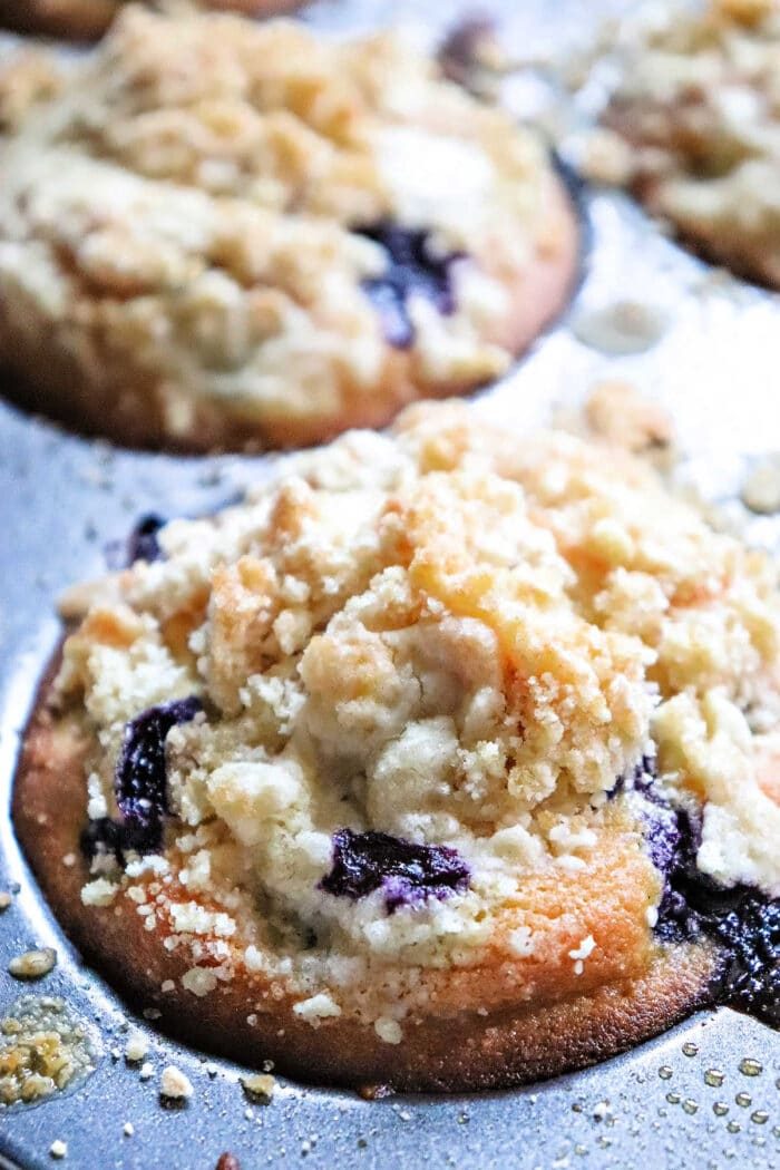A close-up of a blueberry muffin with crumbly streusel topping in a metal muffin pan, showing how Blueberry muffin mix can improve texture for even better muffins.