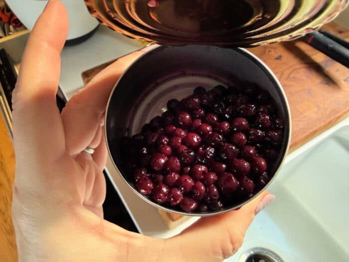 A hand holds an open can filled with dark purple berries over a kitchen sink and cutting board—solid for learning how to make blueberry muffin mix better.