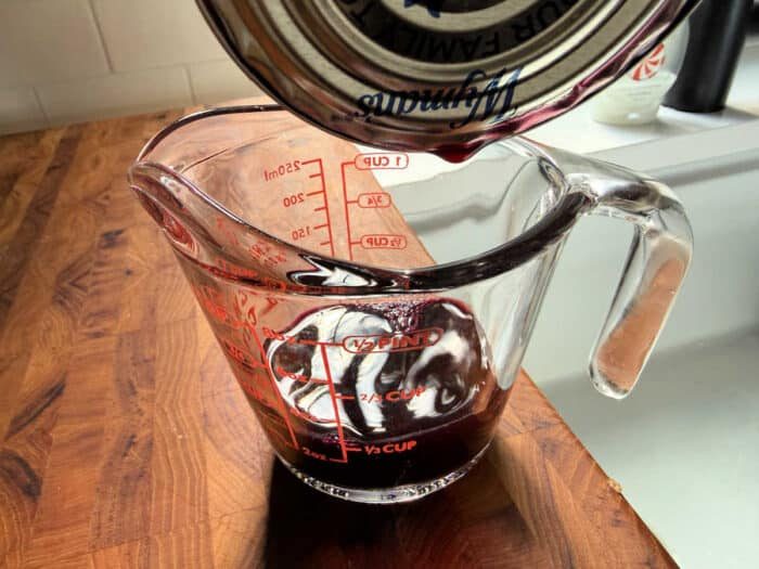 Red liquid being poured from a can into a glass measuring cup on a wooden countertop—solid for learning how to make blueberry muffin mix better with flavorful additions.