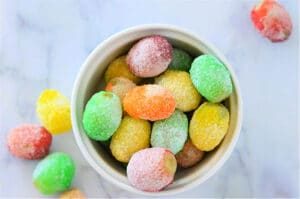 A bowl of colorful, sugar-coated gummy candies on a marble surface sits next to a guide on how to make candied grapes with Jello, adding an extra burst of creativity and flavor to your sweet treats.