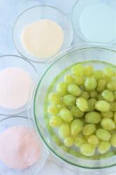 A bowl of green grapes, waiting to be transformed into candied delights with Jello, is surrounded by bowls of white, pink, peach, and cream-colored powders on a marble surface.