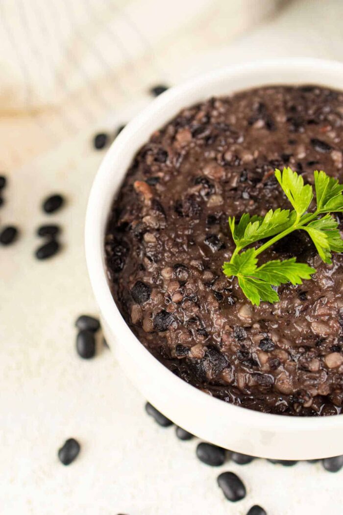 A white bowl of flavorful mashed black bean garnished with parsley is surrounded by scattered black beans, offering an enticing idea on how to make canned refried beans better by blending them into this rich creation.