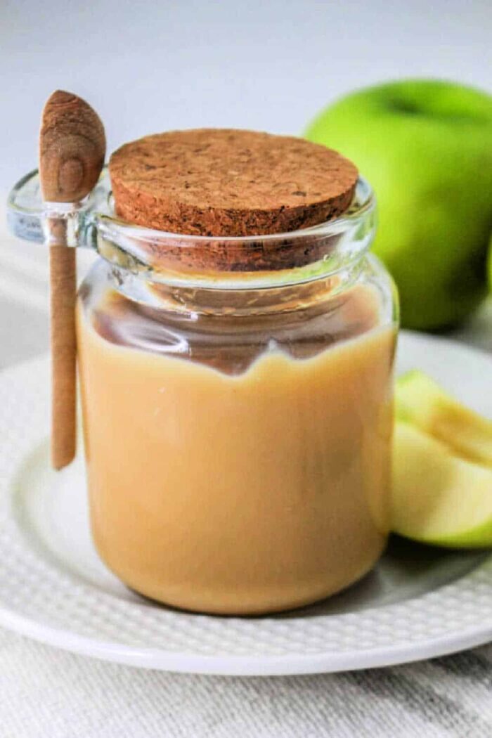 Glass jar of caramel sauce made with condensed milk, cork lid, and a small wooden spoon, next to apple slices on a white plate—solid for those learning how to make caramel with condensed milk at home.