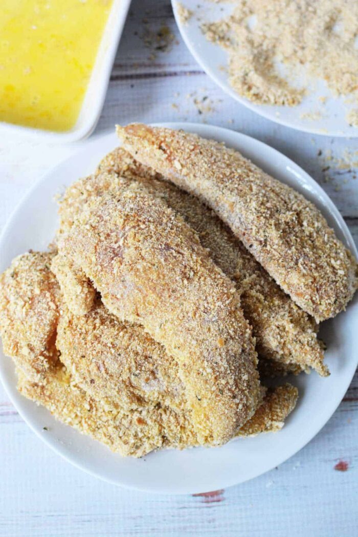 A white plate showcases crispy breaded chicken fillets, while in the backdrop, you can see all the essentials for creating your own masterpiece. This setup might inspire your next cooking adventure after learning how to make chicken tenders at home.