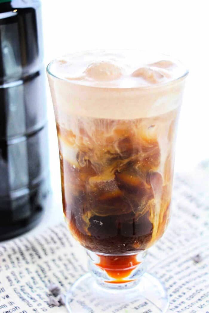 A glass filled with iced coffee topped with frothy cream sits on a surface covered with a newspaper, illustrating how to make cold foam for coffee. The drink has a layered appearance, showing dark coffee at the bottom and swirling milk throughout. A black bottle is partially visible in the background.