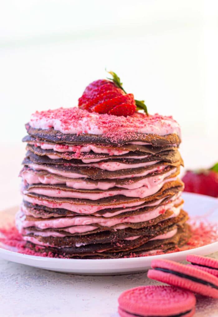 A stack of chocolate crepes layered with pink cream, topped with a strawberry on a white plate, offers an effortless treat reminiscent of learning how to make crepes with pancake mix for elegant brunches.