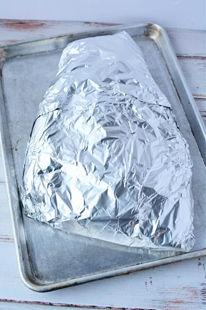 A foil-wrapped brisket in the oven, rests on a baking sheet placed on a white wooden surface.