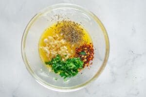 A glass bowl with melted butter, chopped garlic, parsley, red pepper flakes, and Italian herbs on a white surface—what you need for learning How to Make Garlic Bread From Scratch.
