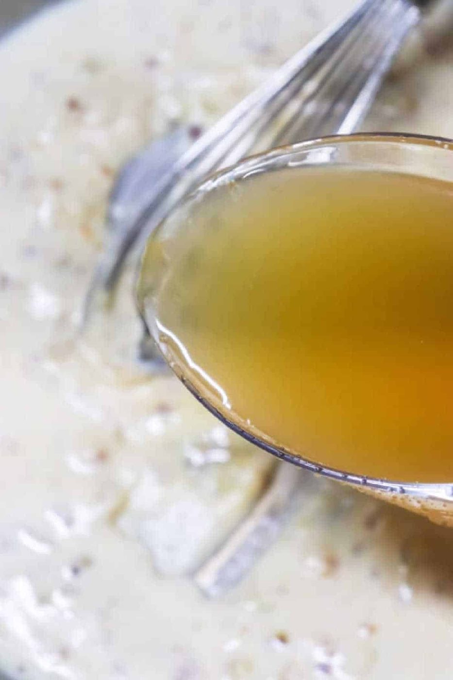 A spoon with clear broth held above creamy soup with a whisk in the background, solid for illustrating how to make gravy from chicken drippings.