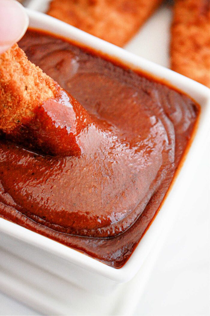 chicken being dipped into rich, popeyes barbecue sauce in a white bowl