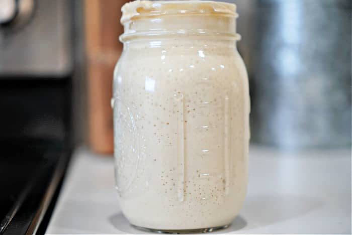 A glass jar filled with creamy, light beige sourdough starter, reminiscent of the process of how to make sourdough starter from scratch, sits elegantly on a kitchen counter.