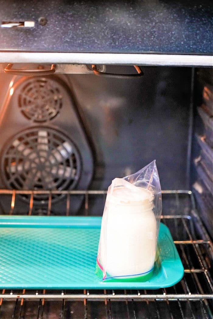 A glass jar sits in a plastic bag on a tray inside an oven, with a fan visible in the back. solid for those learning how to make sourdough starter from scratch, this method maintains the right environment for your homemade fermentation journey.