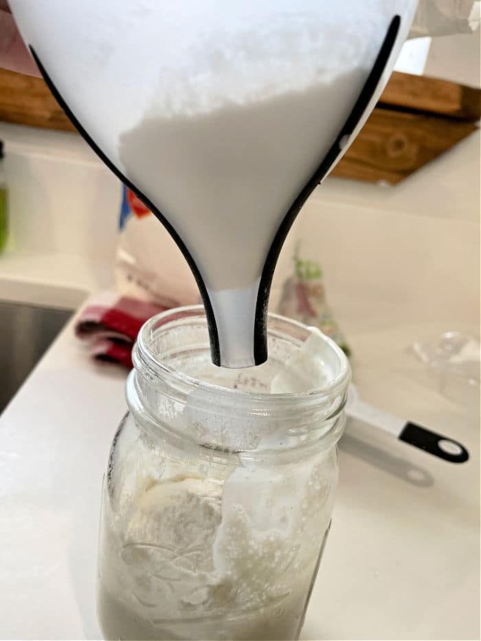 Pouring flour through a funnel into a glass jar on a kitchen counter is the first step in learning how to make sourdough starter from scratch.