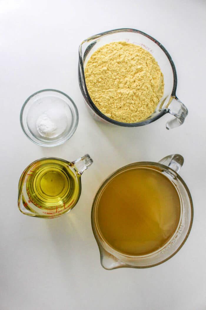 Four measuring cups filled with cornmeal, oil, broth, and salt are arranged on a white surface, showcasing the essential ingredients for how to make tamales masa.