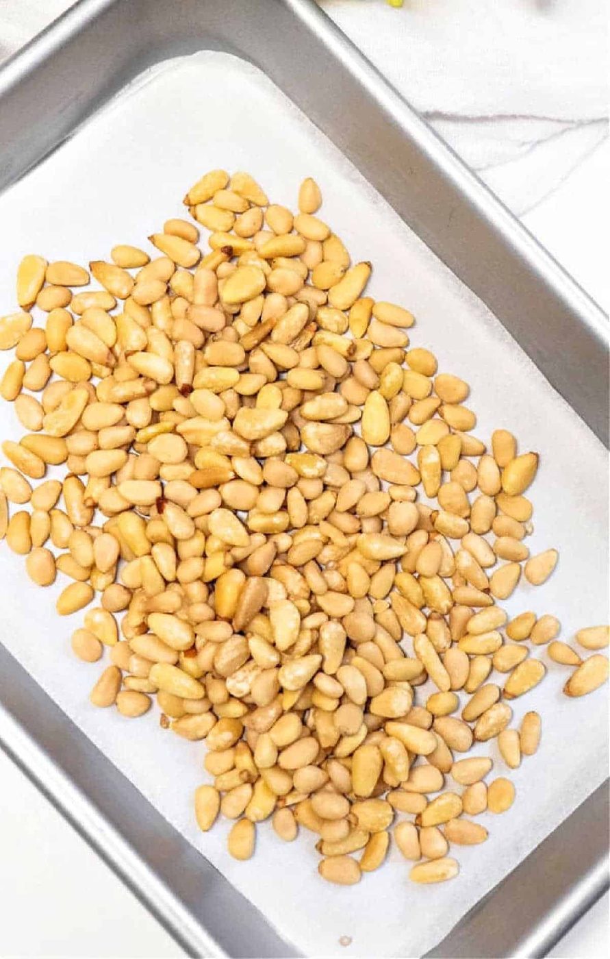 A pile of raw pine nuts spread on a parchment-lined baking tray, ready for roasting—an essential first step in learning how to roast pine nuts to perfection.