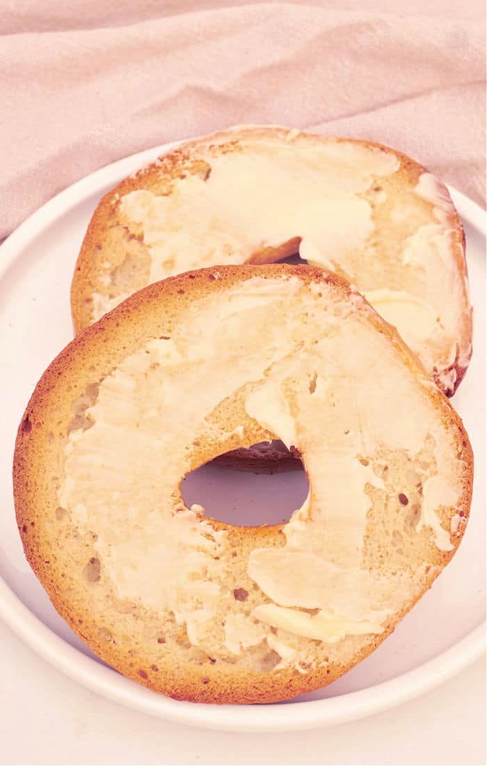 Two buttered bagel halves rest on a white plate atop a light-colored fabric background, tempting you to learn how to toast a bagel in the oven to perfection.