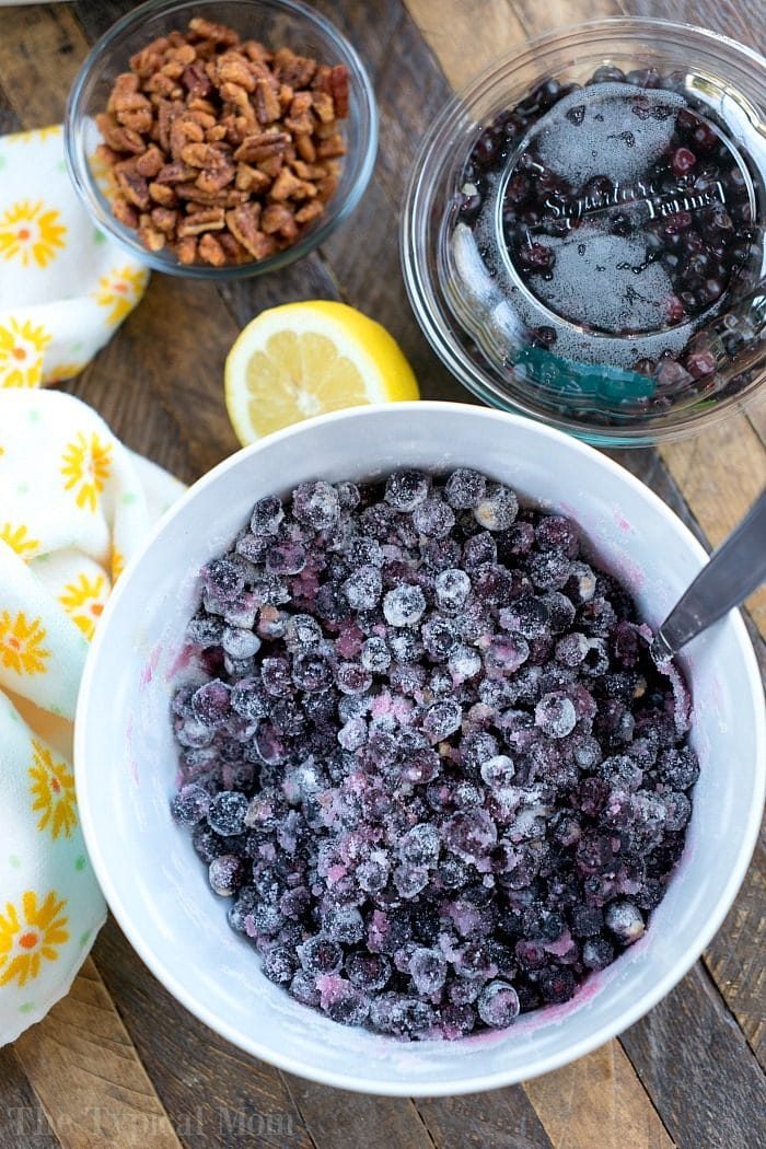 A bowl of fresh huckleberries with a spoon sits next to a lemon, pecans, and more blueberries on a wooden table—solid ingredients for a good huckleberry cobbler.