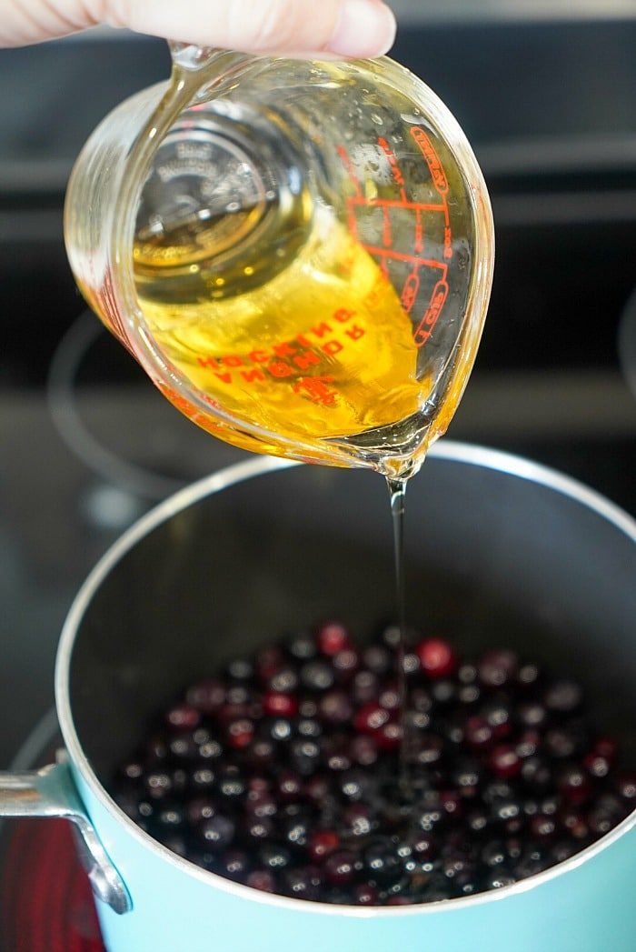 Pouring honey into a saucepan filled with huckleberries on a stovetop.