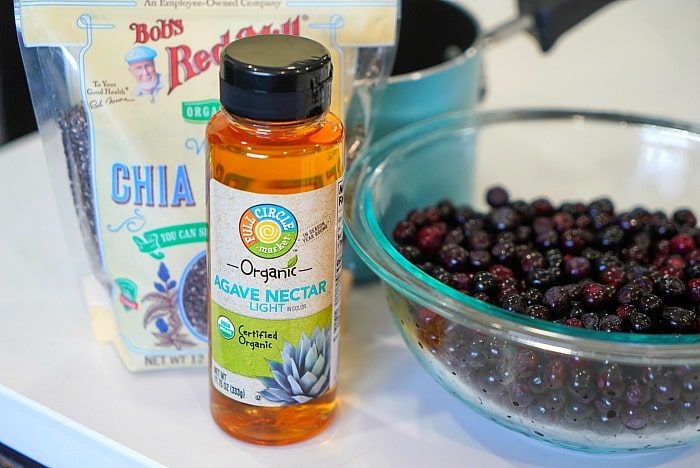 A bottle of agave nectar, a bag of chia seeds, a bowl of fresh berries, and a jar of huckleberry jam sit on the kitchen counter.